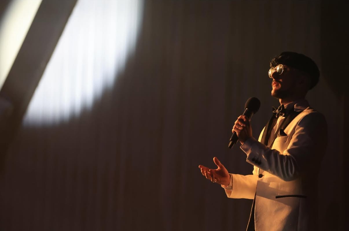 Jake Essex on stage in a cream tuxedo, singing into the light